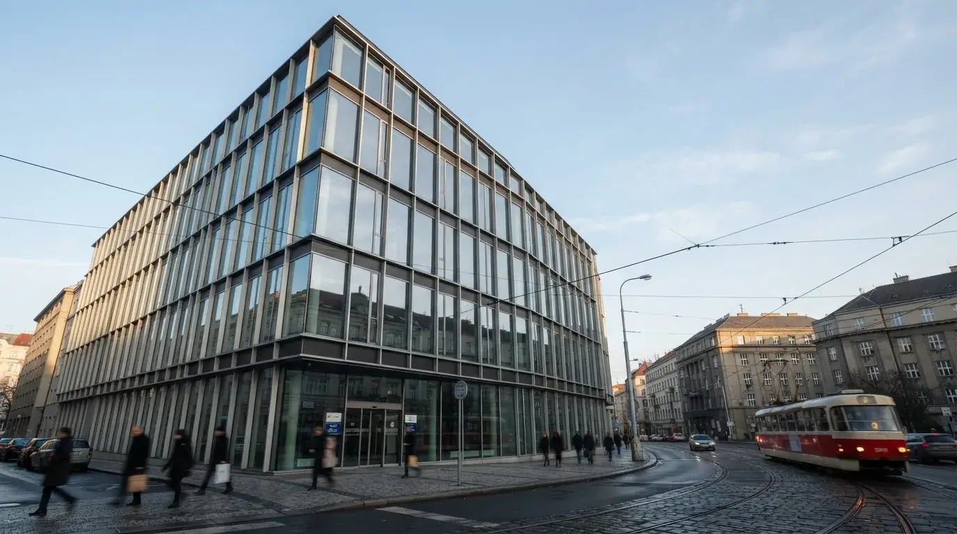 Fasáda moderní administrativní budovy s skleněnými okny odráží ranní oblohu nad rušnou pražskou ulicí.
